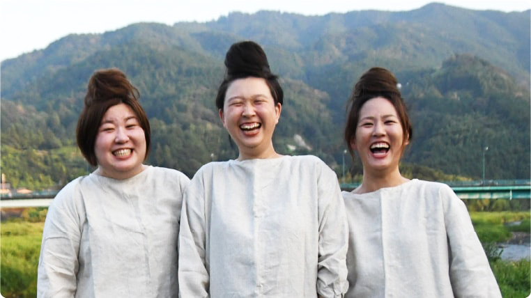 豊かな自然を背景に三人の女性が笑顔で立っている